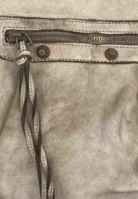 Détail d'un sac en cuir gris avec fermeture éclair dotée d'une tirette tressée, rivets métalliques et surface texturée. Coutures de finition visibles.