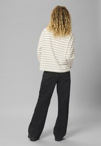 Osoba s kovrčavom plavom kosom, u prugastoj majici dugih rukava i crnim širokim trapericama, stoji okrenuta leđima na jednoličnoj pozadini.