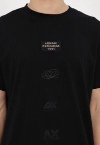 Juodas trumpomis rankovėmis marškinėliai su trimis reljefiniais logotipais: „Armani Exchange 1991“ aplikacija ir „AX“ bei „AM“ grafika žemiau. Lygi audinio tekstūra.