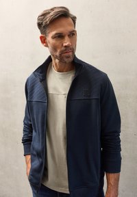 Granatowa kurtka na zamek z panelami o fakturze, zestawiona z jasnobeżową koszulką z okrągłym dekoltem. Model stoi na tle neutralnego betonowego tła.