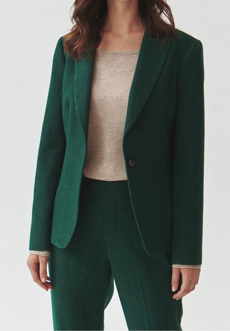 Femme portant un blazer vert foncé et un pantalon assorti par-dessus un top beige à manches longues, debout devant un fond uni.
