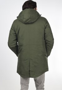 Mørkegrønn isolert parka med stor hette, glidelås foran og sidelommer. Har en rett snitt og en glatt, matt overflate.