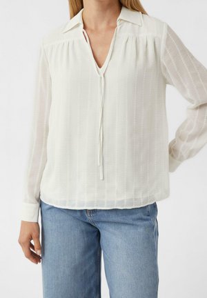 Kvinde iført en hvid langærmet bluse med krave og bindedetalje, kombineret med lyseblå jeans, står med den ene hånd på hoften.