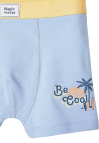 Nahaufnahme von blauen und gelben Shorts mit dem Label "Magic Waves" und dem Text "Be Cool", begleitet von einer Grafik mit einer Palme, einem Sonnenuntergang und Surfboards.