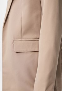 Gros plan sur le tissu beige d'un blazer montrant le rabat de la poche latérale et la manche avec des détails de couture visibles.