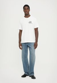 Tricou alb din bumbac cu mâneci scurte, având un grafism negru în stil scris de mână. Asortat cu blugi din denim de culoare albastră deschisă.
