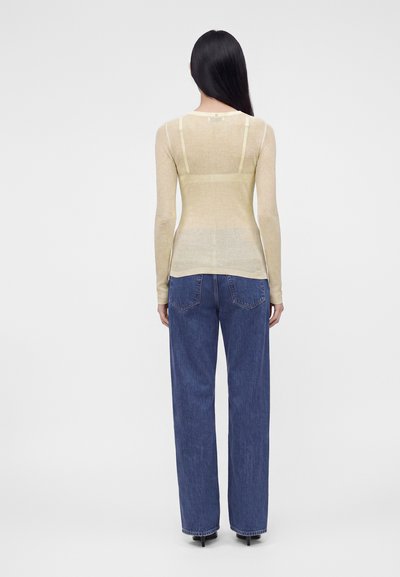 Femme aux longs cheveux noirs portant un haut beige transparent à manches longues et un jean bleu droit taille haute, vue de dos.