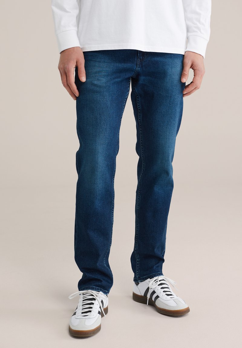 Muž oblečený v modrých úzkych džínsach, bielej košeli s dlhým rukávom a bielych teniskách s čiernymi pruhmi stojaci na jednofarebnom pozadí.