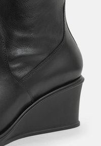 Botte compensée en cuir noir avec une texture lisse, un bout arrondi et un talon angulé distinct. Les détails de couture ajoutent un intérêt visuel.