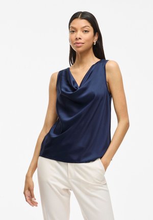 Satijnen mouwloze top in marineblauw met een gedrapeerde voorkant en V-hals. Soepele textuur met een ontspannen pasvorm, gecombineerd met lichtgekleurde broeken.