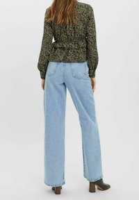 Květinová halenka s dlouhým rukávem v tmavě zelené barvě, kombinovaná se světle modrými vysokým pasem a širokými nohavicemi džín, nošena s černými botami na podpatku.