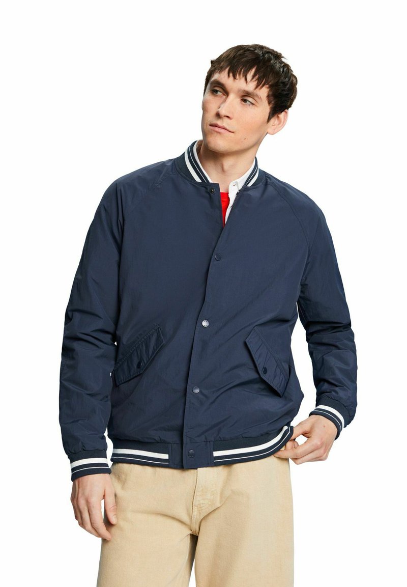 Veste de bombardier marine avec col et poignets côtelés à rayures blanches, dotée de poches latérales et d'un bouton de fermeture. Tissu lisse et léger.