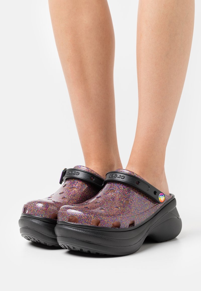 Crocs à plateforme violet pailleté avec des sangles noires et un petit détail circulaire arc-en-ciel, portés pieds nus sur un fond blanc uni.