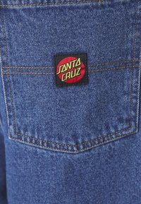 Τζιν ύφασμα σε μπλε απόχρωση με υφή, που διαθέτει μια μαύρη ορθογώνια ετικέτα με το κόκκινο και κίτρινο λογότυπο "SANTA CRUZ" πάνω από μια τσέπη.