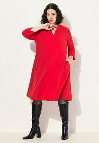 Vestido rojo, de longitud hasta la rodilla, con escote en V y mangas de 3/4. Confeccionado en tejido suave, cuenta con bolsillos laterales. Combinado con botas negras brillantes y de tacón alto.