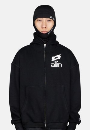 Sweat à capuche zippé noir avec un col montant, doté d'une grande poche avant et d'un logo blanc sur la poitrine et le poignet. Tissu texturé.