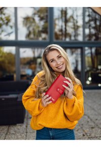 Red leather wallet with a button closure, featuring embossed detail. Model wears a bright yellow sweater and blue jeans, outdoors.