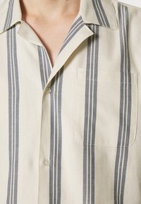 Camisa beige de manga corta con botones en la parte delantera, que presenta rayas verticales azul marino, cuello tipo solapa y detalle de bolsillo en el pecho izquierdo. Textura de tela de algodón.