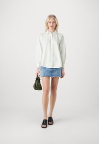 Biała koszula na guziki, dżinsowa spódnica mini z postrzępionym dołem, czarne klapki, zielona torebka. Prosty, czysty design z luźnym krojem.