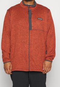Giacca in pile rossa con zip frontale, accenti grigi su colletto e tasche, e un motivo testurizzato in tutto il capo. Logo visibile sul petto.