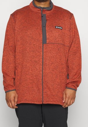 Chaqueta de forro polar roja con cierre frontal, detalles grises en el cuello y los bolsillos, y un patrón texturizado en toda su superficie. Logo mostrado en el pecho.