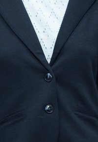 Κοντινό πλάνο ενός μαύρου σακακιού με δύο κουμπιά πάνω σε ένα λευκό υφαντό τοπ με οφθαλμοειδές σχέδιο.