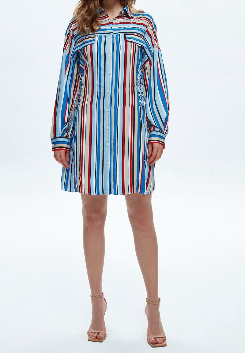 Femme portant une robe à manches longues, col en chemise, boutonnée, avec des rayures verticales bleues, blanches, rouges et noires, et des talons nus à bride à la cheville.