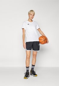 Balts sporta T-krekls ar melnu logotipu, melnas šortus ar baltu apdari un melnus sporta apavus ar dzelteniem akcentiem; tur rokās oranžu basketbola bumbu.