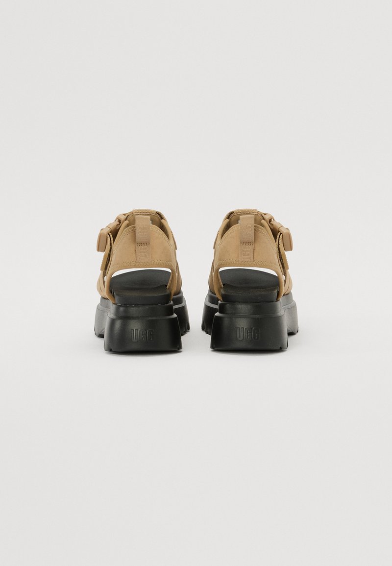 Beige Wildledersandalen mit einer robusten schwarzen Sohle, Riemen an der Rückseite und verstellbaren Schnallen. Sie verfügen über eine glatte Textur und ein minimalistisches Design.