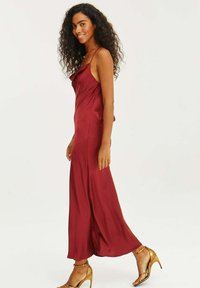 Bordová saténová maxi šaty s výstrihom s kocúrom a tenkými ramienkami, s bočným rozparkom. V kombinácii so zlatými sandálmi na vysokom podpätku. Hladká textúra.