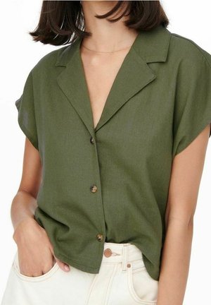 Femme portant une chemise verte à manches courtes avec col, légèrement rentrée dans un pantalon taille haute écru, main dans la poche.