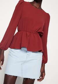 Rød tekstureret bluse med lange ærmer, elastisk taljebånd og udvidet kant, kombineret med en lyseblå denim mininederdel med rå kant og foran syning.