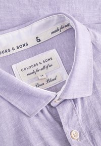 Camicia in misto lino lavanda chiaro con colletto a bottone e cuciture bianche, con etichette che mostrano dettagli sul marchio e sui materiali.