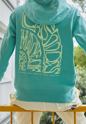 Osoba w turkusowej bluzie z dużym, stylizowanym beżowym napisem na plecach, siedząca na żółtej barierce na zewnątrz, w pobliżu zielonej roślinności i ściany budynku.