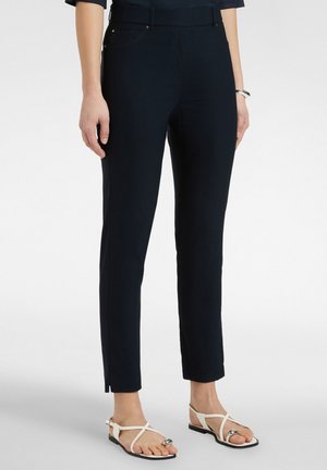 Kobieta w czarnych, dopasowanych, krótkich spodniach i białych płaskich sandałach z pierścionkami na palcach, stojąca na jasnym, jednolitym tle.