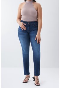 Mujer con jeans azules de talle alto, top sin mangas tipo cuello alto beige y sandalias negras de tacón con tiras, de pie contra un fondo blanco.