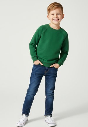 Uśmiechający się młody chłopiec w zielonej bluzie, niebieskich dżinsach i białych trampkach stoi z rękami w kieszeniach na jednolitym tle.