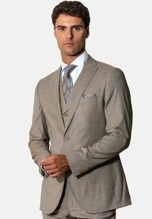 Homme portant un costume trois pièces beige avec une chemise blanche, une cravate et une pochette à motif cachemire, debout les mains jointes.