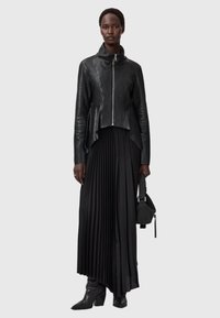 Schwarze Lederjacke mit Reißverschluss und hohem Kragen, kombiniert mit einem schwarzen plissierten Rock und schwarzen Absatzstiefeln, während sie eine kleine strukturierte Tasche hält.