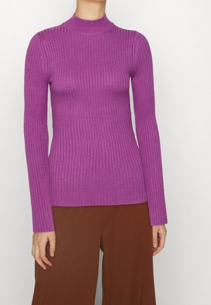 Woman wearing a fitted ribbed purple turtleneck sweater and loose brown pants, standing against a plain light background.