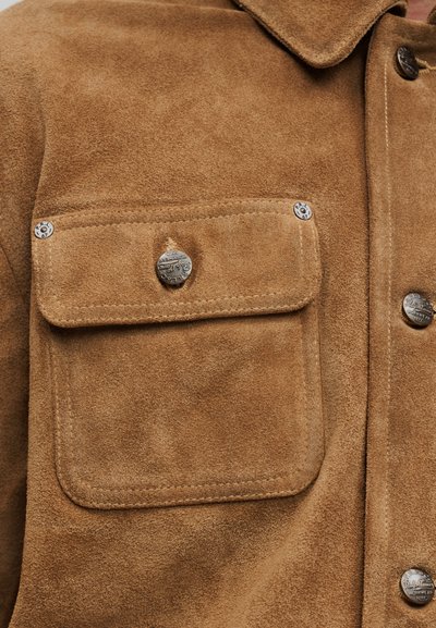 Καφέ πουκάμισο από σουέτ με γιακά, δύο εμπρόσθιες τσέπες με κλεισίματα από ασημένια κουμπιά και εμφανείς ραφές.