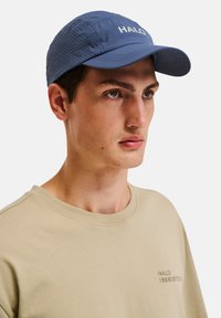 Niebieska czapka z perforowanymi bokami, z białym logo "HALO". Miękka tkanina z zaokrąglonym daszkiem, noszona z prostą beżową koszulą.