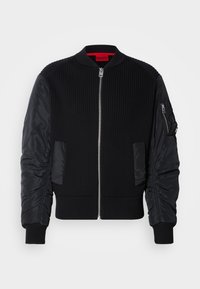 Casaco bomber preto com corpo em malha canelada e mangas lisas de nylon, fecho de correr frontal e bolsos laterais. Corte ajustado com bainhas com punhos.