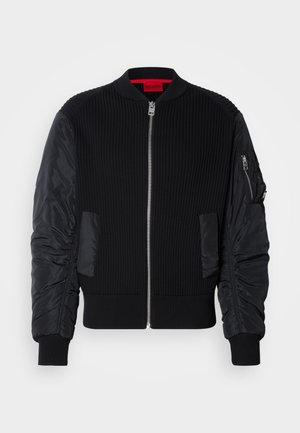 Zwarte bomberjack met geribbelde gebreide body en gladde nylon mouwen, met een ritssluiting aan de voorkant en zijzakken. Getailleerde pasvorm met omgeslagen zoom.