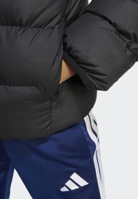 Giacca trapuntata nera con una texture liscia, caratterizzata da un colletto alto e imbottitura visibile. Pantaloni blu navy con strisce bianche adidas e logo.