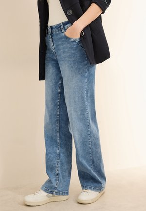 Person iført løstsiddende blå jeans, hvide sneakers, hvid skjorte og sort jakke med den ene hånd i lommen, stående på beige gulv.