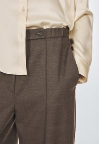 Hnědé tailorované kalhoty s texturovanou tkaninou, které mají elastický pas a boční kapsu. Kombinované se světlou béžovou košilí.