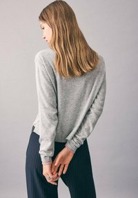 Maglione grigio a maglia con polsini e orlo a costine, con fessure laterali. Abbinato a pantaloni navy a righe sottili, mani incrociate dietro la schiena.