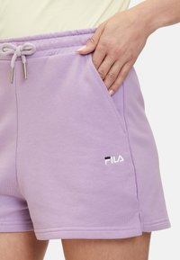 Main droite dans la poche d'un short à cordon lilas FILA porté avec un haut crème, gros plan montrant la texture et le logo sur le tissu.