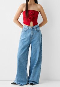 Roter trägerloser Oberteil mit zwei Stoffrosen-Akzenten, kombiniert mit hellblauen, übergroßen Jeans. Die Jeans verfügen über ein klassisches Fünf-Taschen-Design.